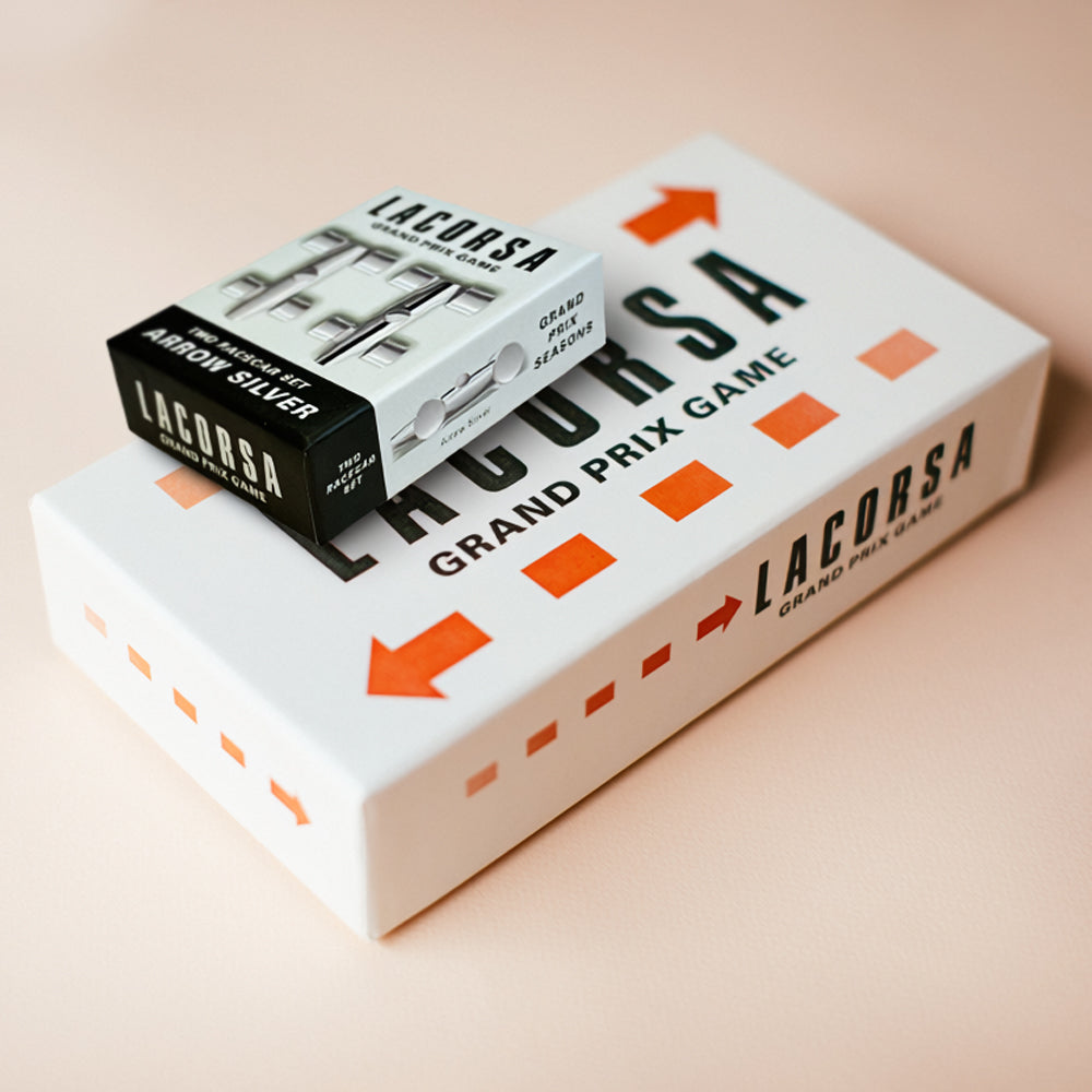 A Lacorsa Grand Prix Game box in white with orange arrows and text, with a smaller black-and-white Arrow Silver box placed on top, photographed on a soft peach background.